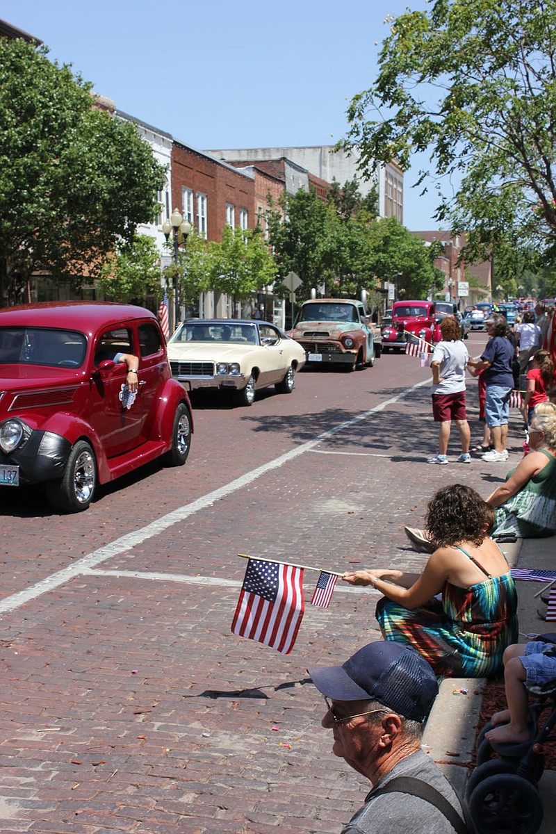 A precession of classic American cars rolls down Court Street during the 4th of July Parade while spectators flag them on.