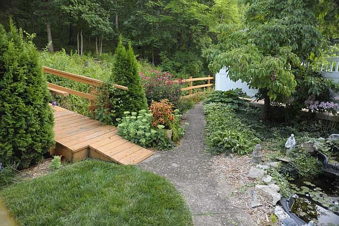 The August Yard of the Month boasts well-cared-for perennials tucked among major features, including a wooden fence line, a bridge spanning a creek bed, an ever-changing fish pond, and a large deck and patio.