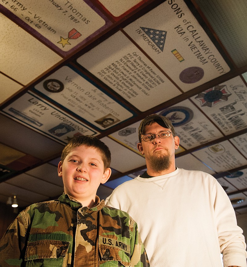 Saxon Scheidegger (left) and his dad, Shawn Scheidegger, stand in the Fulton VFW underneath Shawn Scheidegger's artwork - the ceiling tiles. The tiles commemorate a veteran, alive or lost. Scheidegger has been pursuing art since childhood and said when he enters the Fulton VFW, he "spends the entire time looking up at (the ceiling tiles)." Saxon Scheidegger, 11, said his favorite ceiling tiles are the ones that feature military machinery.