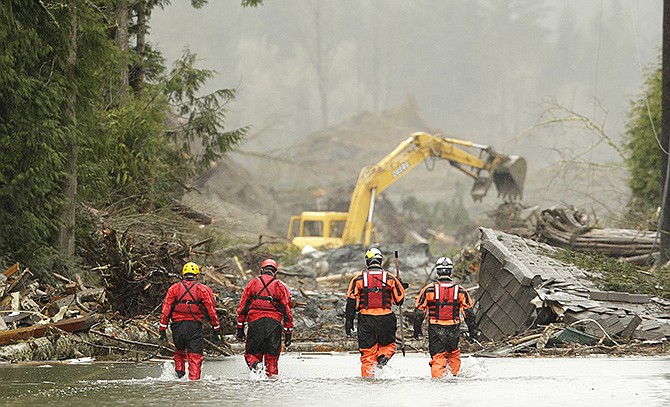 Four search and rescue workers wade Thursday through water covering Washington Highway 530 on the eastern edge of the massive mudslide that struck Saturday near Darrington, Wash., as heavy equipment moves trees and other debris in the background.