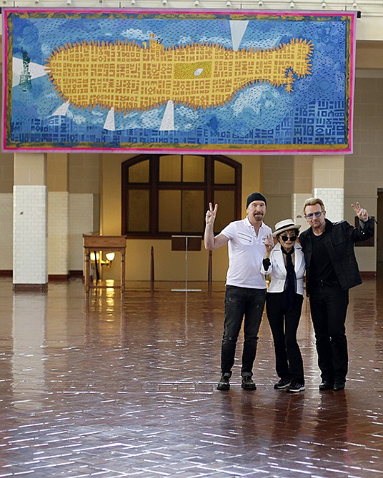 Bono, right, and The Edge, left, of the Irish rock band U2, flank Yoko Ono, the widow of John Lennon, at the unveiling of a giant tapestry depicting the island of Manhattan as a yellow submarine piloted by a waving Lennon at Ellis Island on July 29, 2015 in New York.  