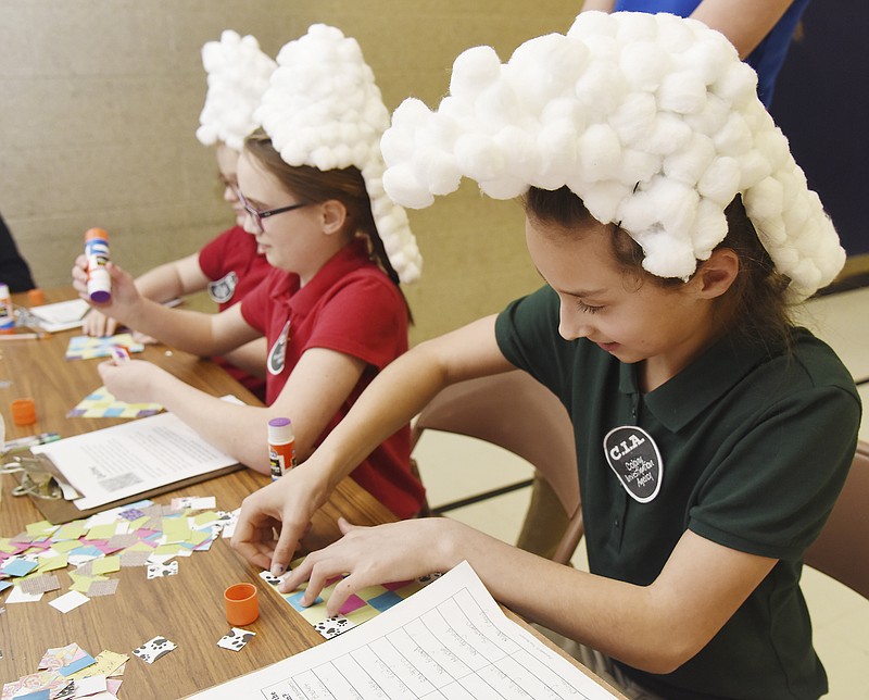 Wearing wigs made of cotton balls glued to white lunch bags, fifth-grade students at Trinity Lutheran School participate in Colonial Days learning stations Wednesday. At the "quilt station," students glued small paper squares onto a piece of construction paper to make a paper quilt. Seated from right, Maddie Homfeldt, Lauren Reid and Kate Bailey. 