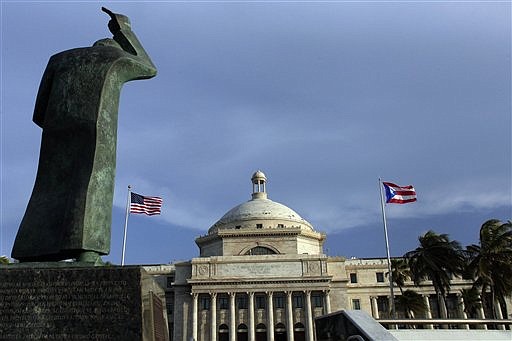 In this Wednesday, July 29, 2015, file photo, a bronze statue of San Juan Bautista stands in front of Puerto Rico's Capitol as U.S. and Puerto Rican flags fly in San Juan, Puerto Rico.
