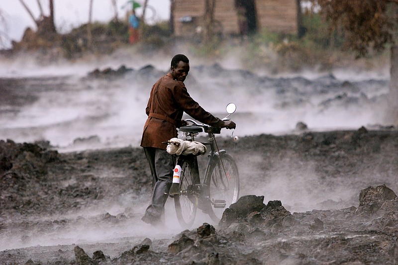 In this file photo taken Thursday, Jan. 24, 2002, a Congolese man pushes his bicycle across lava rock covered in steam, after a rain storm in the eastern Congolese town of Goma. Traumatized farmers are slowly returning to fields decimated by the 2002 eruption of Mount Nyiragongo in eastern Congo. Flowing lava flattened more than 30 percent of the city of Goma, 20 kilometers away. 