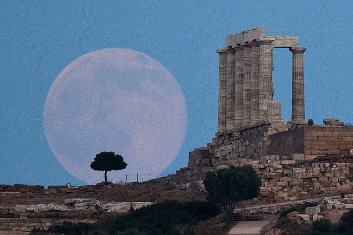 A full moon rises behind a tgree nextg to the ruins of the ancient marble Temple of Poseidon, built in 444 BC, southeast of Athens, Greece. 