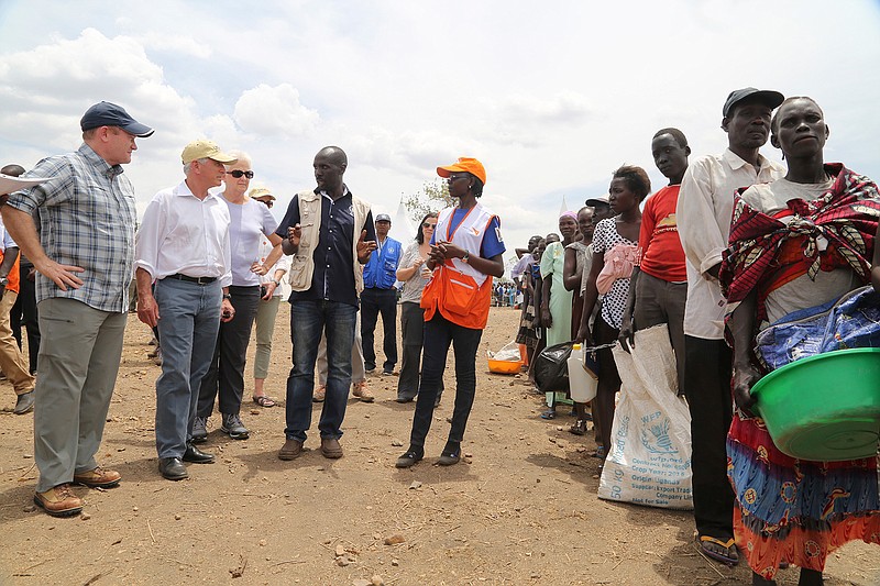 In this photo taken on Friday April 14, 2017, U.S Senators Chris Coons, left, and Bob Corker, second left, listen to an aid worker as South Sudanese refugees wait for a food distribution at the Bidi Bidi refugee settlement in northern Uganda. In a political climate dominated by President Donald Trump's slogan of "America First," two U.S. senators are proposing making American food aid more efficient after meeting with victims of South Sudan's famine and civil war. After visiting the world's largest refugee settlement in northern Uganda, Democratic Sen. Chris Coons of Delaware told The Associated Press that the U.S. "can deliver more food aid at less cost" through foreign food aid reform.