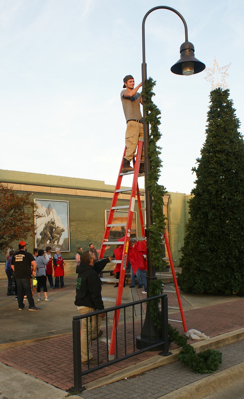 Kenneth Parnell of Freeman's Landscaping in Texarkana is high on the ladder placing wreaths on the 111 lamp posts Nov. 9 in downtown Atlanta, Texas, while in the background Atlanta Area Chamber of Commerce members prepares for a holiday photograph.