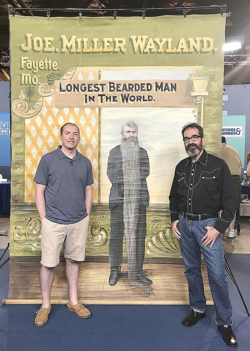 Michael Kuster, left, poses in front of a banner featuring an image of his great-great-great-grandfather, Joe Miller Wayland. The poster was commissioned in 1901. Antiques Roadshow appraiser Gary Piattoni is shown at right. 