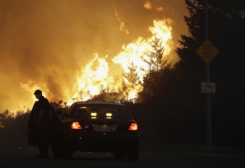 FILE - In this Monday, Oct. 9, 2017, file photo, a law enforcement officer blocks a road as flames burn in a residential area in Santa Rosa, Calif. Police body-camera footage from 2017's wildfires in California's wine country shows officers running door-to-door urging people to flee and rescuing elderly residents of a retirement community as flames bear down. Video obtained by the San Jose Mercury News is from the point-of-view of police in Santa Rosa, as they sprint through swirling smoke amid the firestorm. (AP Photo/Jeff Chiu, File)