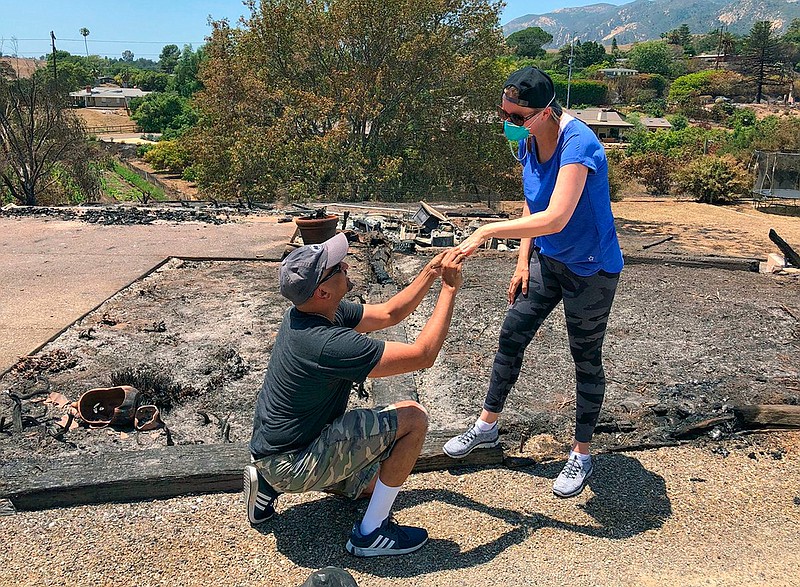 In this photo provided by the Santa Barbara County Fire Department resident Ishu Rao on one knee, places this wife's wedding ring on her finger, next to the charred remains of their home in Goleta, Calif., Sunday, July 8, 2018.  The California couple who lost their home in a wildfire made a new happy memory amid the ashes when they found what was left of Laura Rao's wedding ring. Ishu and Laura Rao returned to the rubble of their home of three years on Sunday, to look for her Tiffany ring. They searched with the help of some firefighters in the area and Ishu Rao made the find, prompting him to get down on one knee and again propose to his bride. (Mike Eliason/Santa Barbara County Fire Department via AP)