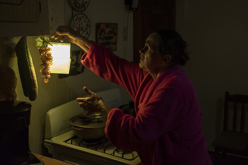 In this July 12, 2018 photo, Marta Bermudez Robles, 66, hangs a lamp in her kitchen in Adjuntas, Puerto Rico, at her home that is still with electricity since Hurricane Irma and Maria. The only power Bermudez and her husband have had for 10 months is courtesy of a neighbor who threw over an extension cord connected to his generator that provides just enough power to light one bulb in the kitchen and another in the living room for a couple hours each day. (AP Photo/Dennis M. Rivera Pichardo)