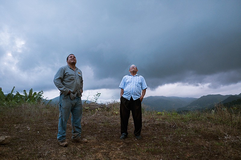 Retired resident Ramon Serrano, right, stands outside his home with a lineman from the Puerto Rico Power Authority as they watch a man restore power that was cut off by hurricanes Irma and Maria in Adjuntas, Puerto Rico. For the first time in 10 months, Serrano watched the 11 o'clock news on a recent weeknight and went to bed at midnight with his wife. "It's the latest we've been up," the 77-year-old said.