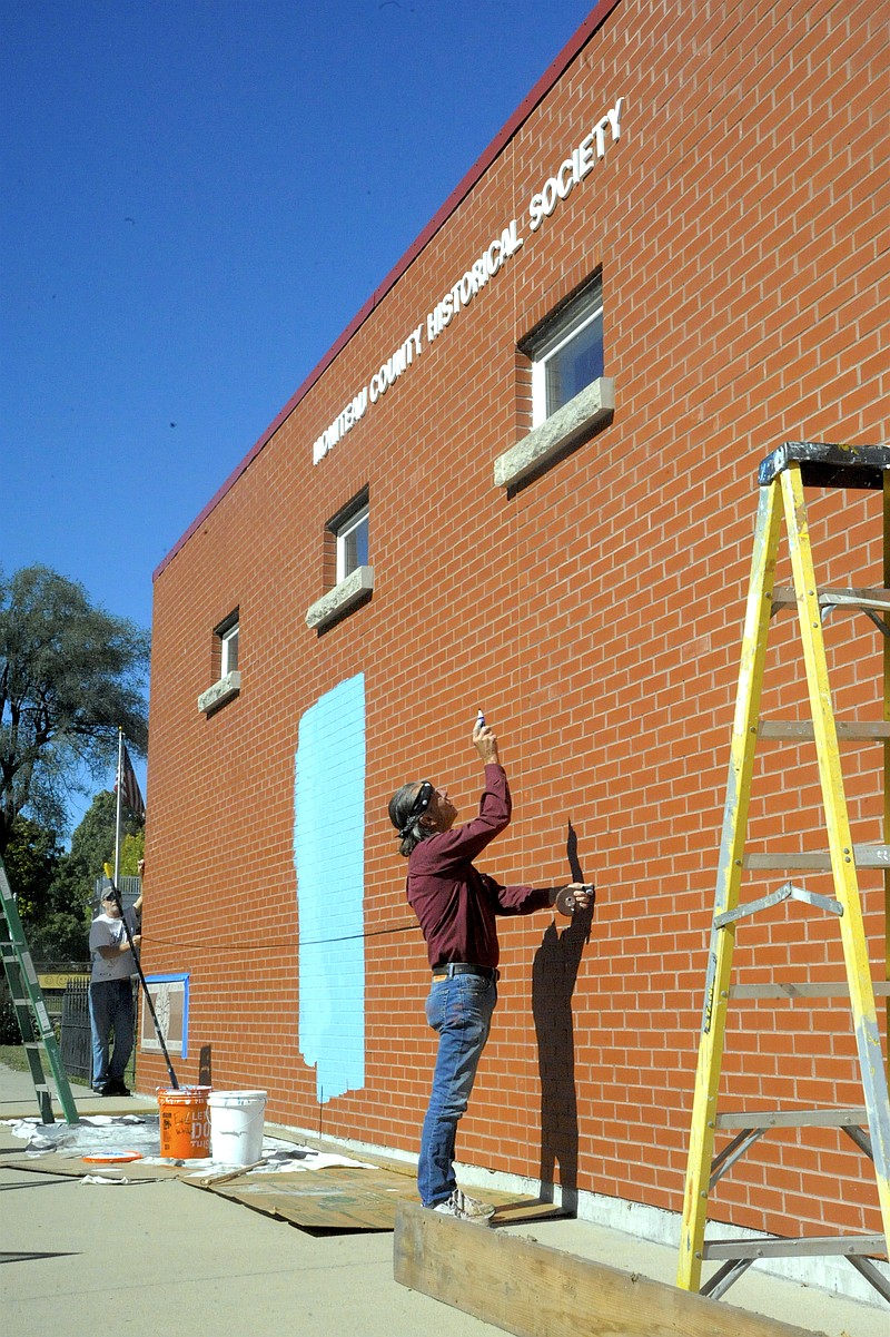Muralist Dennis Holliday measures for the center of his 10x60-feet mural, highlighting scenes at the Moniteau County Fair, on the Moniteau County Historical Society's genealogy library wall facing South Street with the help of Danny Bentzen.