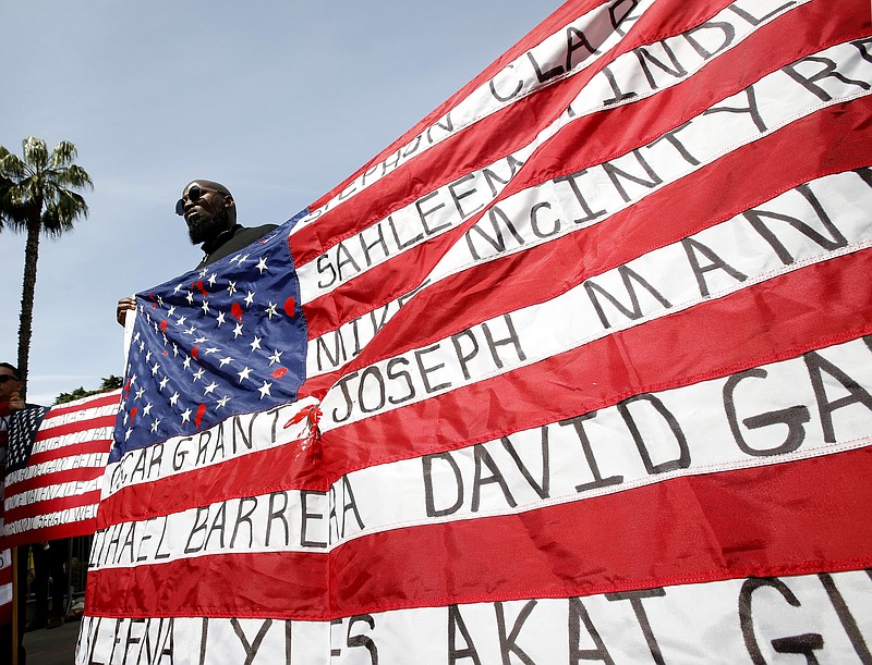 File - In this April 8, 2019, file photo, Malaki Seku Amen holds up an American flag with the names of people shot and killed by law enforcement officers, as he joins other in support of a bill that would restrict the use of deadly force by police in Sacramento, Calif. Round two is under way in California's fight over how best to limit fatal shootings by police through nation-leading reforms. State legislators on Tuesday, April 23, 2019, are debating a measure that proponents say would set a national precedent by creating statewide guidelines on when officers can use lethal force and requiring that every officer be trained in ways to avoid opening fire. (AP Photo/Rich Pedroncelli, File)