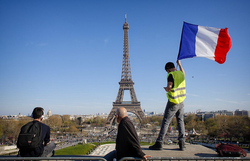 FILE - In this Saturday, March 30, 2019 file photo, a man holds a French flag as French with the Eiffel Tower in the background, during protests in Paris. Paris is wishing the Eiffel Tower a happy birthday with an elaborate laser show retracing the monument's 130-year history. First, the monument invited 1,300 children to a giant "snack time" Wednesday, May 15 beneath the tower dubbed the Iron Lady. (AP Photo/Thibault Camus, file)