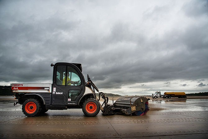 Under a sky of dark clouds, Natasha Faulkner, who works for the Parking Division, operates a Bobcat UTV 4x4 with a rotary brush attachment to clean the tarmac near the terminal at Jefferson City Memorial Airport. She was part of a crew of staff from Jefferson City Public Works, Parking Division and MoDOT who worked cooperatively Friday to remove mud caked on the pavement. Now that the water has receded for the second time from much of the paved areas, the cleanup can begin again. 