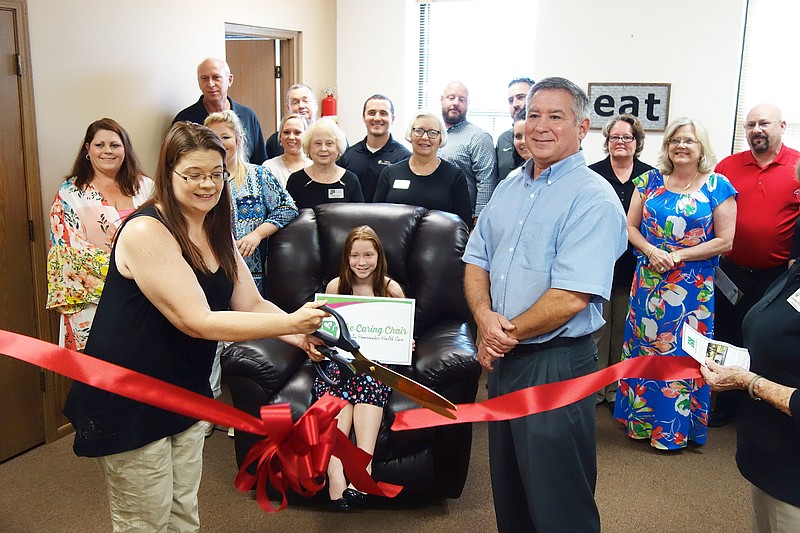 Sarah Wood, left, and Jeff Buker of Homemaker Health Care cut a ribbon while members of the Callaway Chamber of Commerce look on. HHC announced its newest program Wednesday named The Caring Chair. Meredith Buker, center, is perched in the chair.