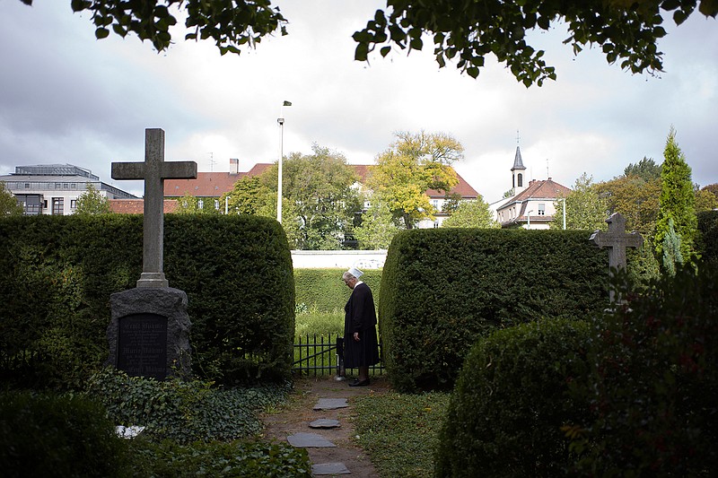 In this Sept. 18, 2019, photo, Sister Brigitte Queisser of the Lutheran Lazarus Order stands near graves of the Lazarus sisters during an interview with The Associated Press in Berlin. For many years, the deaconesses lived in West Berlin across the street from the Berlin Wall in a mother house that is seen in the background. The sisters' order ran a hospital where many of the eastern escapees were treated. (AP Photo/Markus Schreiber)