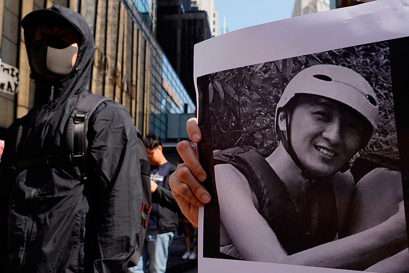 A protester stands near a photo of Chow Tsz-Lok during a memorial flash mob to remember him in Hong Kong on Friday, Nov. 8, 2019. Chow, a Hong Kong university student who fell off a parking garage after police fired tear gas during clashes with anti-government protesters died Friday, in a rare fatality after five months of unrest that intensified anger in the semi-autonomous Chinese territory. (AP Photo/Vincent Yu)