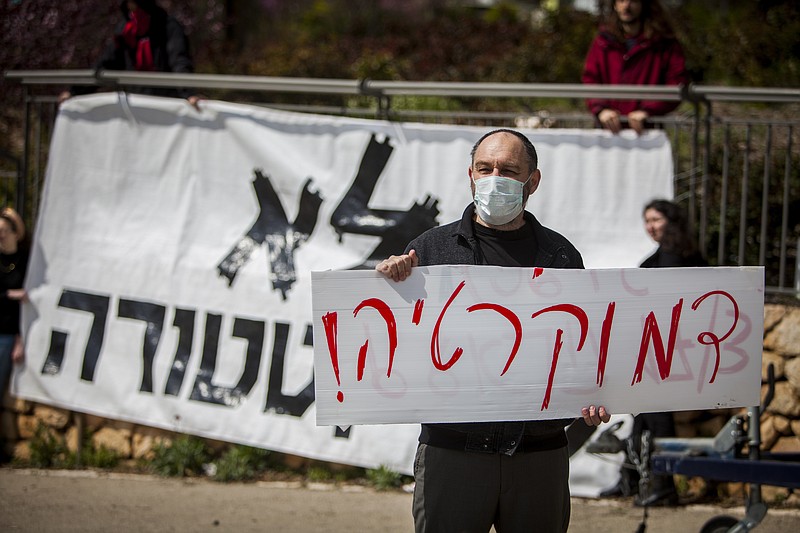 A man holds a sign that reads in Hebrew; "Democracy" during a protest outside the Israeli parliament in Jerusalem, Thursday, March 19, 2020. Hundreds of people defied restrictions on large gatherings to protest outside parliament Thursday, while scores of others were blocked by police from reaching the area as they accused Prime Minister Benjamin Netanyahu's government of exploiting the coronavirus crisis to solidify his power and undermine Israel's democratic foundations. (AP Photo/Eyal Warshavsky)