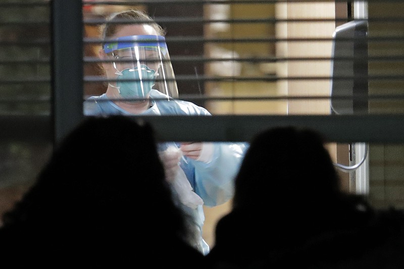 FILE - In this March 10, 2020, file photo, a worker wearing protective gear is seen through a window as she works in a room of a woman who has tested positive for the new coronavirus, as her daughters look in from outside the window, at the Life Care Center in Kirkland, Wash., near Seattle. Burgeoning coronavirus outbreaks at this and other nursing homes in Illinois, New Jersey and elsewhere are laying bare the risks of the industry’s long-running problems, including a struggle to control infections and a staffing crisis that relies on poorly paid aides who can't afford to stay home sick.  (AP Photo/Ted S. Warren, File)