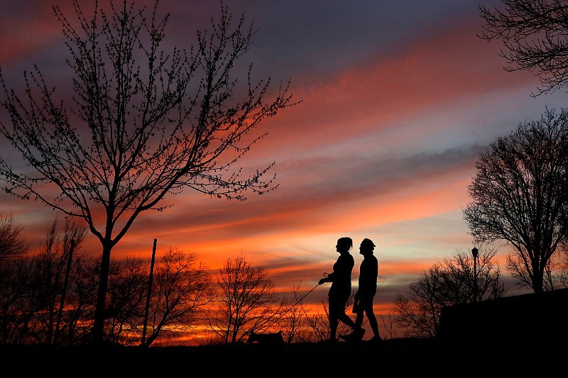 A couple walk a dog at sunset, Wednesday, March 25, 2020, in Kansas City, Mo. The city, along with neighboring counties, is under Stay at Home orders to help prevent the spread of COVID-19, the disease caused by the new coronavirus. (AP Photo/Charlie Riedel)