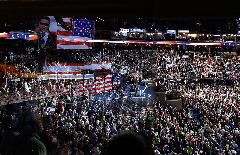 In this July 26, 2016, file photo the stage is reflected on a glass window on the suite level at Wells Fargo Arena as Timmy Kelly sings the national anthem before the start of the second day session of the Democratic National Convention in Philadelphia. The coronavirus pandemic is forcing Democrats and Republicans to take a close look at whether they'll be able to move forward as planned this summer with conventions that typically kick off the general election season. (AP Photo/John Locher, File)