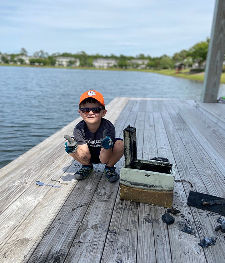 In this May 9, 2020, photo provided by Catherine Brewer, Knox Brewer poses next to a safe he pulled out of Whitney Lake in South Carolina. The 6-year-old was using a magnet attached to a string to fish for metal in the water when he reeled in a lockbox that police said was stolen from a woman who lived nearby eight years ago. (Catherine Brewer via AP)