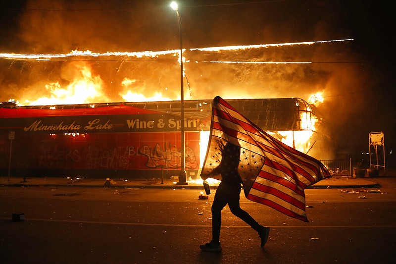 A protester carries a U.S. flag upside, a sign of distress, next to a burning building Thursday, May 28, 2020, in Minneapolis. Protests over the death of George Floyd, a black man who died in police custody Monday, broke out in Minneapolis for a third straight night. (AP Photo/Julio Cortez)