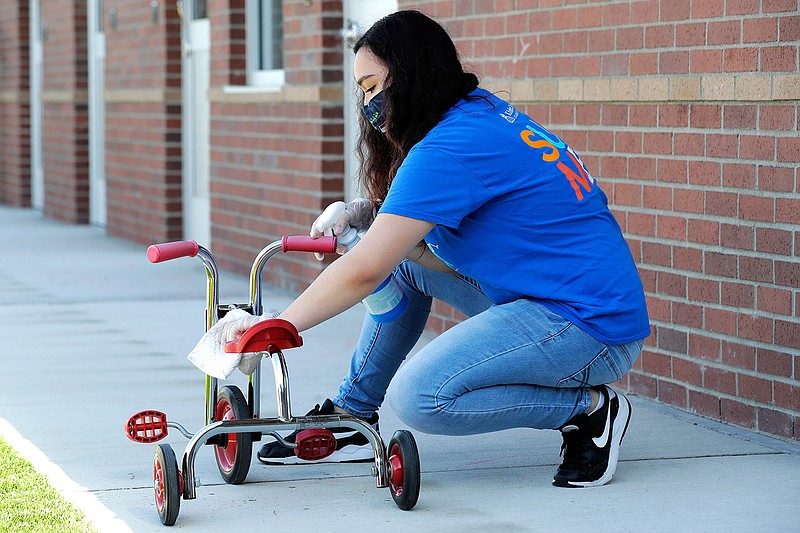 In this May 27, 2020 photo, Alena Kleinman, a worker at the Frederickson KinderCare daycare center in Tacoma, Wash., wears a mask as she cleans a tricycle following use by a class, a task that is repeated several times a day. In a world weary of the coronavirus, many working parents with young children are now struggling with the decision on when or how they'll be comfortable returning to their child care providers. Frederickson KinderCare has been open throughout the pandemic to care for children of essential workers. (AP Photo/Ted S. Warren)