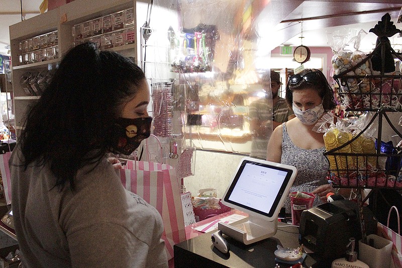 In this Thursday, May 28, 2020, photo, an employee at Bruce's Candy Kitchen ring up a customer's purchases from behind a protective plastic shield as both wear face masks due to the coronavirus in Cannon Beach, Ore. With summer looming, Cannon Beach and thousands of other small, tourist-dependent towns nationwide are struggling to balance fears of contagion with their economic survival in what could be a make-or-break summer. (AP Photo/Gillian Flaccus)