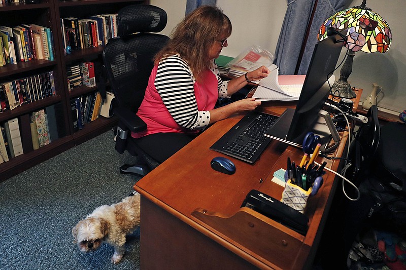 Ellen Boudreau works remotely at her home, Tuesday, Aug. 4, 2020, in Manchester, N.H. Boudreau, who was laid off from her full-time bookkeeping job for six weeks as the COVID-19 virus pandemic shutdown much of the area businesses, now works less than 10 hours per week with that employer, while also taking on a part-time, temporary worker job at another office. Walking out from behind her desk is her 15-year-old dog "Happy Go Lucky". (AP Photo/Charles Krupa)