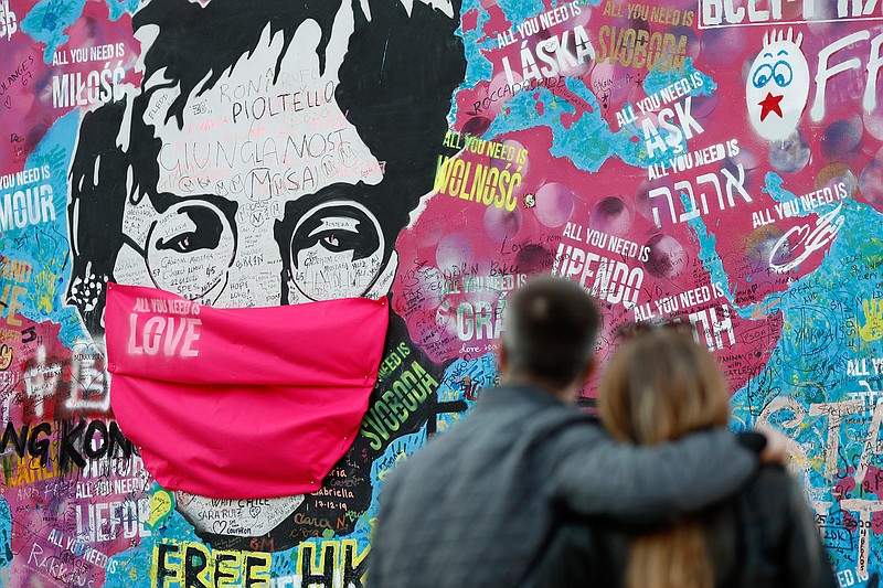 A couple look at the "Lennon Wall" with a face mask attached to the image of John Lennon, in Prague, Czech Republic, on April 6, 2020. Like so many other events in the year of coronavirus, an annual tribute to John Lennon held in its adopted city of New York will go online. The five-hour event will be streamed for free on Lennon's birthday, October 9, starting at 7 p.m. Eastern time on the LennonTribute.org website. It will feature recorded performances from Patti Smith, Rosanne Cash, Natalie Merchant, Jackson Browne, Jorma Kaukonen and others. (AP Photo/Petr David Josek)