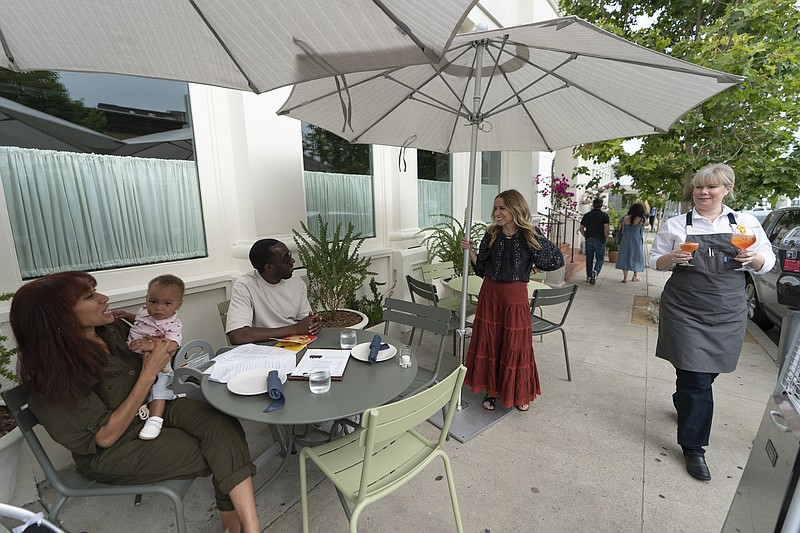 In this Saturday, June 19, 2021, photo, Caroline Styne, owner and wine director at The Lucques Group, standing under umbrella, welcomes back regular customers, Chris Anokute with his wife Jasmine and their 9-month-old son, Phoenix, at the A.O.C. Brentwood restaurant in Los Angeles. Styne has turned away dozens of customers at the company's A.O.C. West Hollywood restaurant because she doesn't have the staff to serve them, leaving seats empty. (AP Photo/Damian Dovarganes)