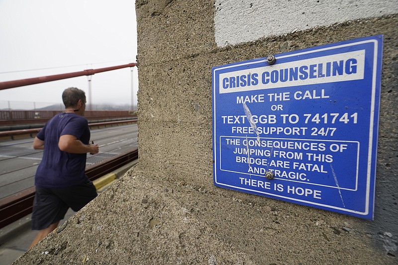 In this Aug. 3, 2021 photo a man jogs past a sign about crisis counseling on the Golden Gate Bridge in San Francisco. In recent years, officials who oversee so-called suicide hotspots like San Francisco’s Golden Gate Bridge and the George Washington Bridge, which spans New York City and New Jersey, have worked to install prevention or deterrent systems. (AP Photo/Eric Risberg)