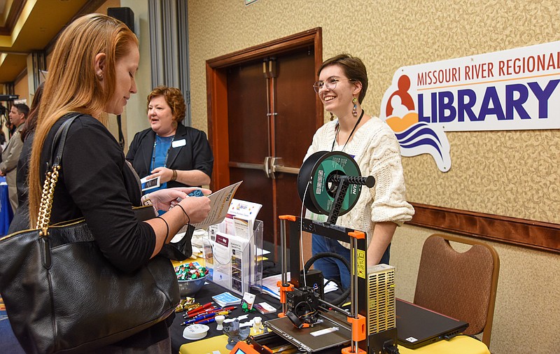 <p>Julie Smith/News Tribune</p><p>Allie Gladbach, right, graphic artist for Missouri River Regional Library, explains to Danielle Briot the library has a 3-D printer, seen in foreground, the public can access. Briot works for the Office Of Equal Opportunity and was visiting the booths at Wednesday’s Business Leadership Summit at Capital Plaza Hotel. In the background is Claudia Cook, director of MRRL.</p>