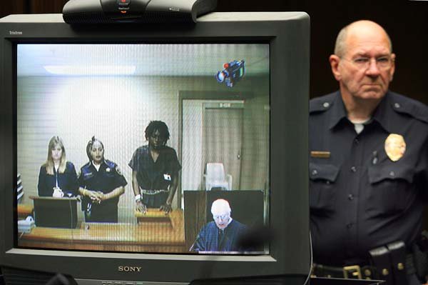 Laquan Javaris Fitzpatrick (center) appears on a video monitor in North Little Rock District Court Thursday morning during his arraignment on capital-murder charges. North Little Rock police officer Wayne Chance (right) looks on as Judge Jim Hamilton (lower right corner of the video screen) takes Fitzpatrick’s plea of innocent. 
