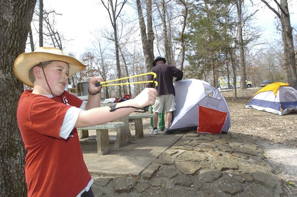 GENERATIONAL GETAWAY J.J. Meyl aims his slingshot Friday while his grandpa, Mike Meyl, sets his tent up in one of the limited off-season campsites at Prairie Creek park on Beaver Lake. All of Prairie Creek park and five other Beaver Lake parks open Friday, followed by all parks opening May 1. J.J., his brother Tyler Meyl and their grandfather, all of Fayetteville, marked the end of spring break with a camping trip.
