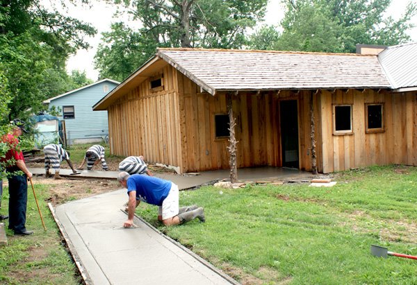 Three inmates of the Benton County Sheriff’s Department inmate work detail recently completed new sidewalks at the entrance of a new exhibit building at the Gravette Historical Museum. The rustic building will be open by Gravette Day the weekend of August 12-13. Shown with the inmates are Steve Mitchael, foreground, who is trowling wet cement, and Leland Brandon. Both are members of the Museum Commission.

