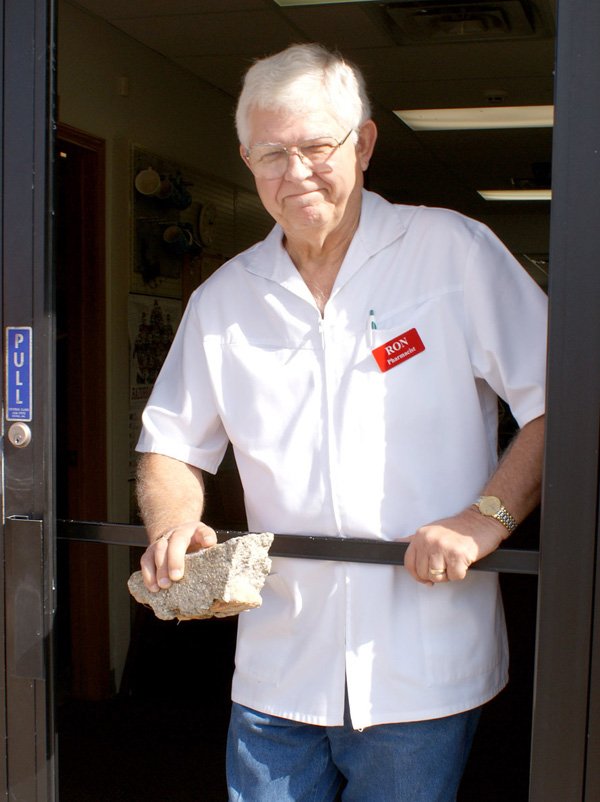 Gravette pharmacist, Ron Teasley, looks out through the frame of what was the back door to Teasley Pharmacy which was burglarized early last Wednesday. Teasley is holding a large concrete/stone, similar to the one used to smash the glass in the store door.