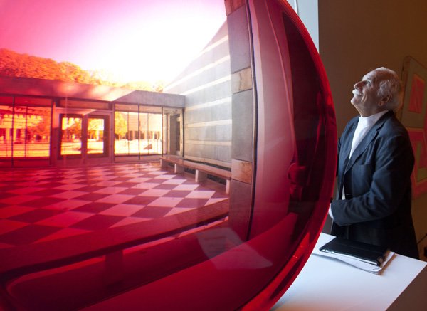 DESIGN DIMENSIONS Architect Moshe Safdie looks out of a window Thursday next to a large red untitled magnifying disk sculpture by artist Fred Eversley at Crystal Bridges Museum of American Art in Bentonville. “I want people to leave the place uplifted, and never forget it. Is that too much to ask?” he said.
