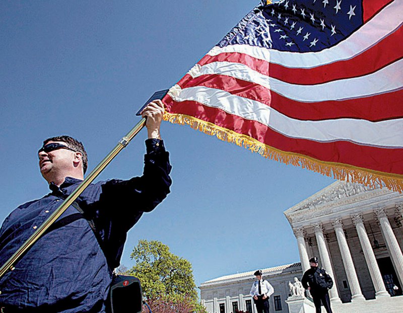 Holding an American flag and a copy of the Constitution, Dan, who is from Virginia and didn’t want his last name used, protests against the health-care law Monday outside of the Supreme Court in Washington after the first day of arguments. 