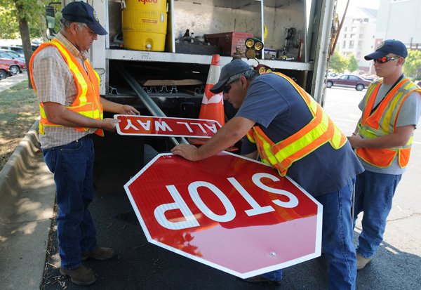 Kole Horne, right, watches as fellow State Highway employees Len Williams, center, and Jerry Howe bolt a new stop sign together Thursday to replace signs and add another stop sign on West Maple Road at the intersection with North Razorback Road. The fourth sign makes the intersection a four-way stop. Stop signs at the intersection were replaced with larger, 36-inch signs. An electric sign has been placed on West Maple Road for 30 days to inform drivers of the change. 