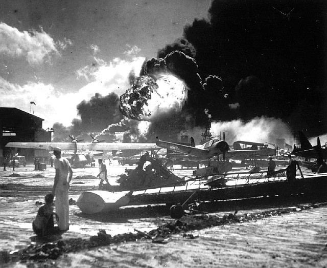 Sailors stand among the wreckage on the airfield at Ford Island Naval Air Station on Dec. 7, 1941, as they watch a ship explode in the background. 