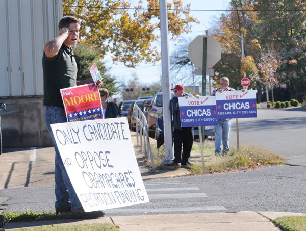 Joel Keller, from left, Mike Carney and Jim Murawski hold political signs Thursday outside of an early voting location in downtown Rogers. Candidates can purchase tickets to events hosted by other candidates, according to Arkansas Ethics Commission campaign rules. Investigations are complaint-driven, and violations can result in letters of caution, warning or reprimand; a fine of $50 to $2,000, or misdemeanor criminal charges. 