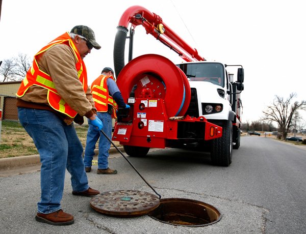 Johnny Lunsford, left, removes the manhole cover while Pat Sharp, both with Rogers Water Utilities, prepares to lower a hose into the sewer line under South Third Street on Friday in Rogers. The Vac-Con system sends water at 60 gallons/minute and 3000 psi of pressure to clean trash and other materials from the sewer line. 