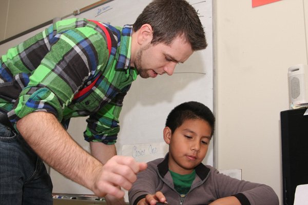 Kyle Queen, fourth-grade teacher at Bonnie Grimes Elementary School, helps Allan Alfaro with computer research during class in Rogers. Queen is one of 39 men employed as elementary school teachers in the Rogers School District. 