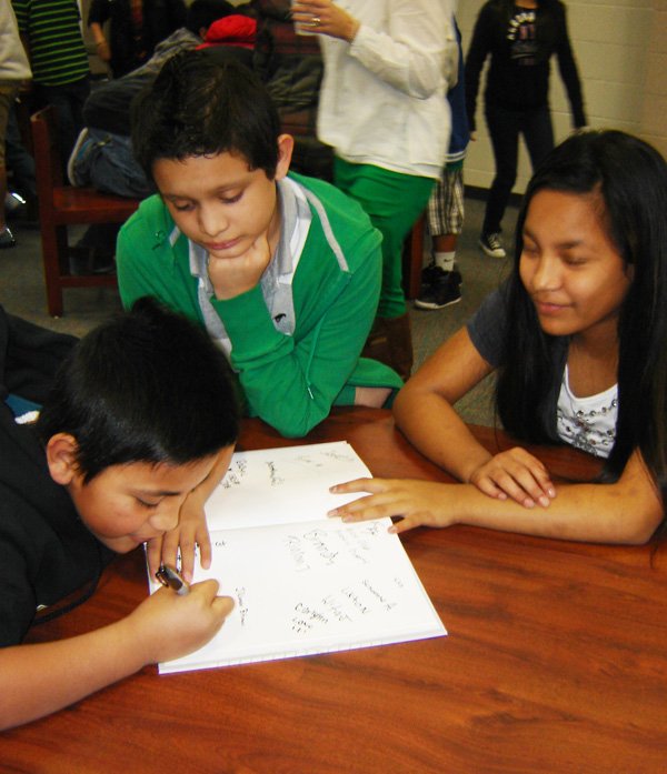 Nambo Konou, left, signs a copy of the book “First Crossing: Kids with ‘Real’ Stories About Immigrating to the United States,” as Jose Mancias and Matilda Carmel look on Monday during a book signing at Sonora Middle School in Springdale.