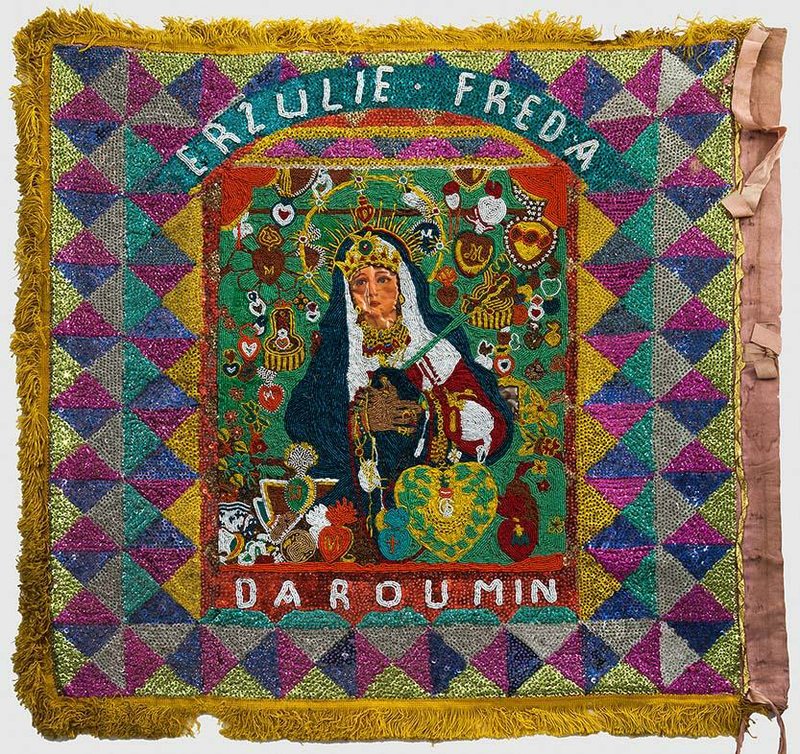 Ezili (Erzulie) Freda Daroumin shows the blend of Catholic and Western African influences. Here, Mary Queen of Sorrows is depicted as Ezili in this flag from the mid-20th century. It is made from fabric, plastic, sequins, beads, ribbon, fringe and chromolithographic image. It is part of “Sacred Symbols in Sequins: Vintage Haitian Vodou Flags” at the University of Arkansas at Little Rock Gallery I. 