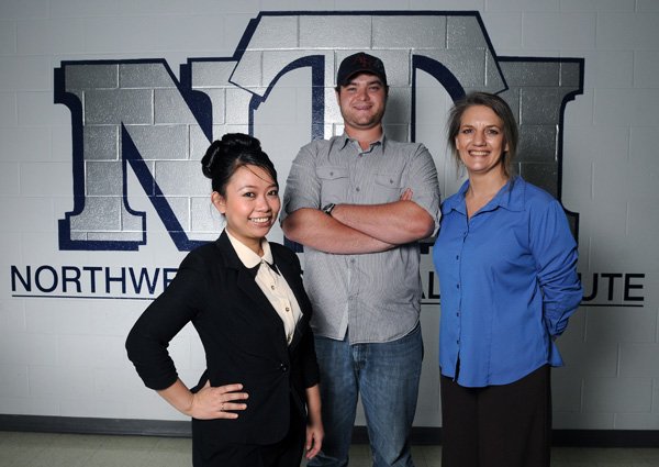 Tonya Carter, from right, Cody Pharris and Chanitnun Hernandez stand in front of a logo for NTI on June 13 at Northwest Technical Institute in Springdale. The students will be getting their general education diploma at the same time as their diplomas from NTI. 